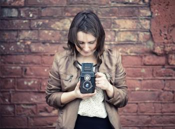 Pretty young woman holding retro camera and taking photo on vintage brick wall background. Selective focus on camera. Toned photo with copy space. Vintage style photo.