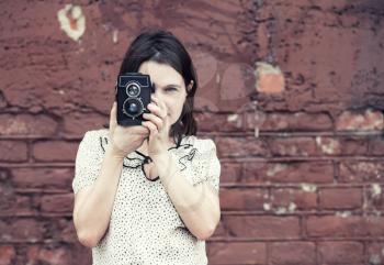 Girl holding retro camera and taking photo on vintage brick wall background. Selective focus on camera and hands of model. Toned photo with copy space. Vintage style photo.