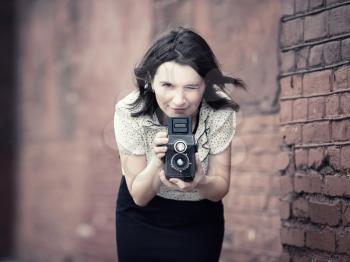 Woman with vintage camera in hands against blurred vintage brick wall background. Pretty young woman taking photo outdoors. Selective focus on the model's face and camera. Vintage style photo.