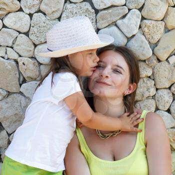 Hugging mother and daughter. Smiling woman and baby girl in white hat. An old stone wall in the background.