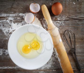 Cooking or baking. Still life with eggs, raw eggs in a dish, eggshells, flour, rolling pin and whisk on wooden background.