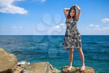 Pretty young woman standing on a stone and posing with his hands behind his head against a background of sea and blue sky. Selective focus on the model.