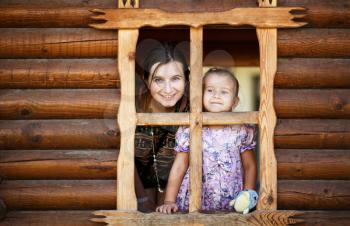 Pretty young mother and daughter a little girl look out of the window a wooden hut. Happy smiling mother and daughter.