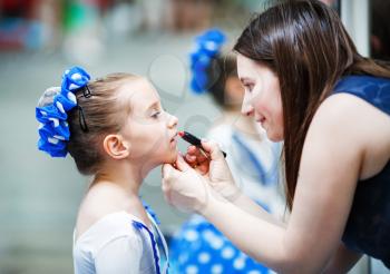 Mother putting lipstick on her daughter on mirror background. Mom helping little daughter to use lipstick before a dance performance. Mom makes make-up her daughter. Shallow depth of field. Selective 