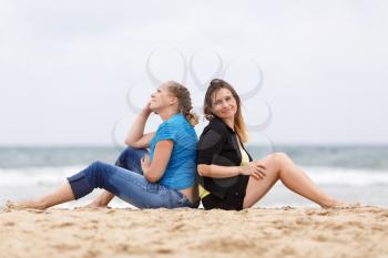 Two pretty young smiling woman sitting and relaxing on the sand on the seashore. Shallow depth of field. Focus on models.