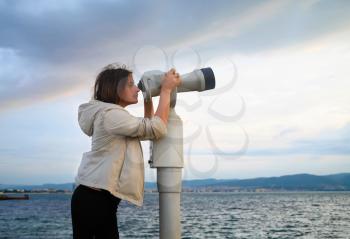 Pretty young woman using a coin operated binocular enjoying a great view of the Black sea. Early morning. Shallow depth of field. Focus on the model.