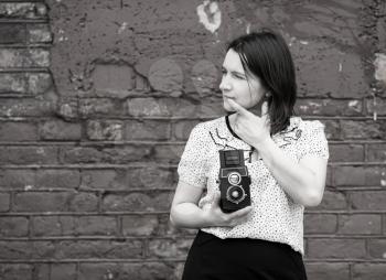 Woman photographer with retro camera in hand. Girl is thinking and looking away. Monochrome image. Old vintage grunge wall in the background. Selective focus on the model.