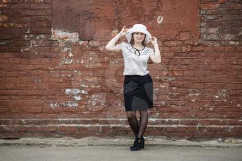 Photo of a woman in a white hat, blouse and black skirt, standing against the backdrop of an old vintage brown brick wall. Model holds the edges of the hat by hand. Portrait of a woman in a retro styl
