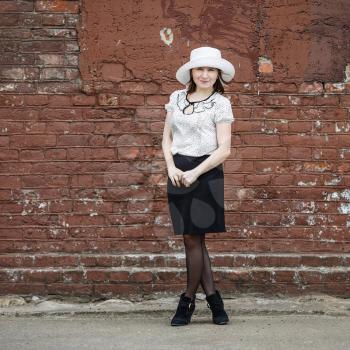 Photo of a woman in a white hat, blouse and black skirt, standing against the backdrop of an old vintage brown brick wall. Space for text.