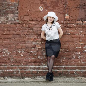 Pretty young brunette woman in a white hat, blouse and black skirt, posing outdoor in old vintage brown brick wall background. Girl is leaning against the wall. Space for text.