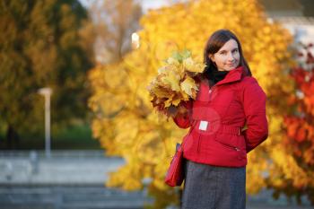 Female portrait against a bright autumn foliage. Young woman in red jacket holding in your hand a yellow maple leaves. Selective focus on model.
