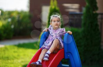 A child in a smart dress and sandals sitting on slide and dreamily looking up. Cute little girl. Warm sunny summer day. Shallow depth of field. Selective focus on the model's face.