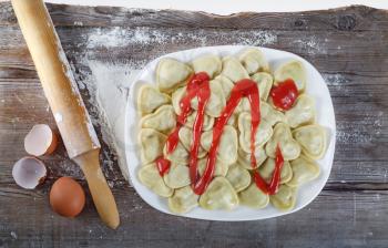 Dish of ravioli with ketchup, a rolling pin, eggs, flour and egg shells on a wooden background. Top view.