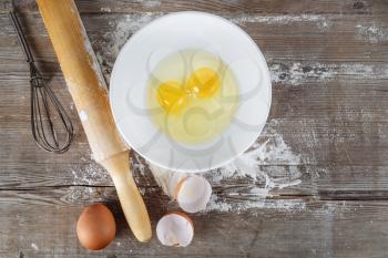 Cooking background with eggs, raw eggs in a dish, eggshells, flour, rolling pin and whisk on a wooden background.