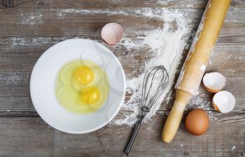 Baking background with eggs, raw eggs in a dish, eggshells, flour, rolling pin and whisk on a wooden background. Top view.
