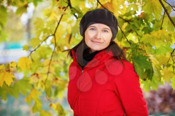Pretty young woman in a beret and red jacket on the background of green and yellow autumn maple leaves. Female portrait. Selective focus on model.