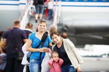 Two young smiling women and child girl posing in the blurry background ladder passenger aircraft at the airport. Passengers on the airliner background. Shallow depth of field. Selective focus.