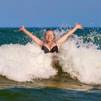 Beautiful smiling girl with her hands raised in the foam of a sea wave. Happy young woman. Bright sunny day.