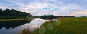 Evening rural landscape. River with thickets of trees and grass on the banks. The calm surface of the water with the reflection of the evening sky. Panoramic shot.