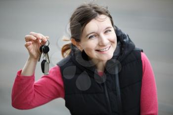 Smiling young woman holding car key on blurred gray asphalt background. Selective focus.