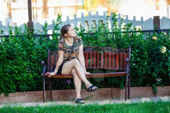 Pretty young woman sitting on a bench in the park on background of bright green foliage. Selective focus on girl.