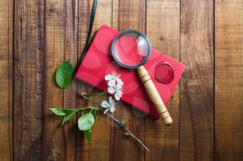 Photo of red notebook, magnifier, wax seal, herry flowers and green leaves on vintage wooden background. Stationery elements.