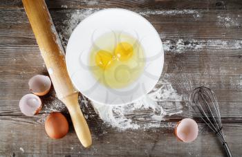 Still life with raw eggs, rolling pin and whisk on vintage wooden table background. Top view.
