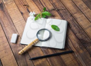 Photo of blank notebook, magnifier, pencil, eraser, cherry flowers and green leaves on wooden background. Mock up for placing your design.
