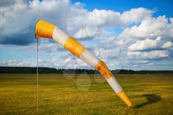 Striped orange and white windsock against blue sky with clouds and field of grass.