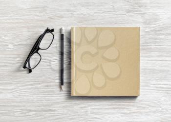 Photo of closed blank square book or notepad, glasses and pencil on wooden background. Template for placing your design. Flat lay.