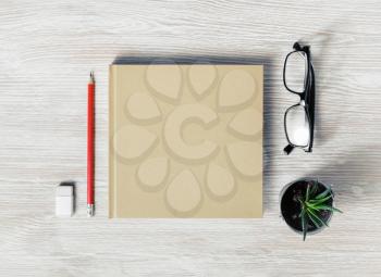Photo of closed blank square booklet and stationery: glasses, pencil, eraser and plant on light wooden background. Template for placing your design. Flat lay.
