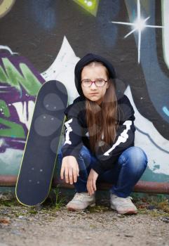 Child girl sitting near a skateboard against the wall. Vertical shot. Selective focus.