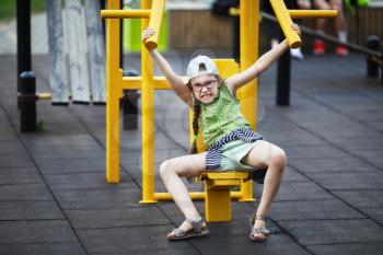Angry little girl making exercises on sports training apparatus outdoors. Child goes in for sports and shakes muscles. Selective focus.