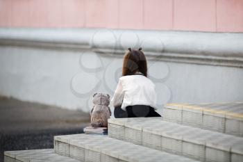 Sad girl with a plush toy sits on the steps of the school stairs with her back to the camera. Offended child. Selective focus.