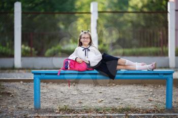 Schoolgirl in school uniform resting on a bench. Smiling child girl leaning on backpack. Selective focus.