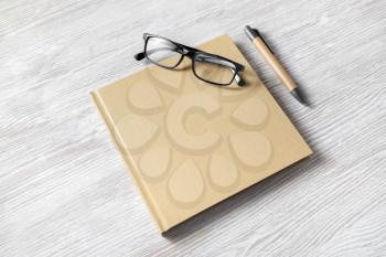 Photo of closed blank square book, glasses and pen on light wooden background. Template for placing your design.