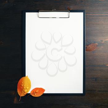 Blank white sheet of paper in clipboard and autumn leaves with water droplets on wood table background. Flat lay.