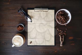 Stationery set and coffee. Clipboard with blank kraft sheet of paper, coffee cup, coffee beans, glasses and ground powder on wooden background. Flat lay.