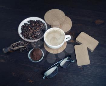 Coffee cup, roasted coffee beans, ground powder, beer coasters, blank kraft business cards and glasses on kitchen table background.
