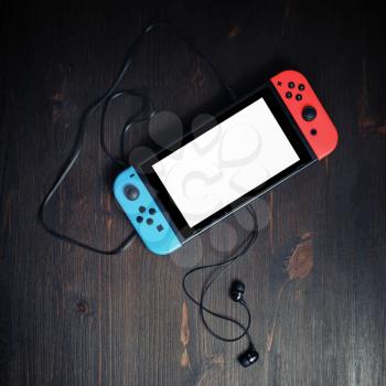Minsk, Belarus - May 07, 2020: Nintendo Switch game console with blank white screen and headphones on vintage wooden background. Flat lay.