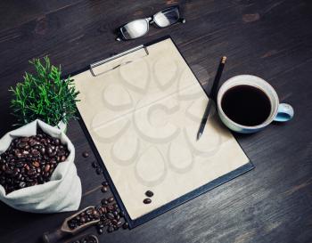 Stationery and coffee. Clipboard with blank kraft paper, coffee cup, plant, coffee beans in canvas bag, glasses and pencil on wooden background.