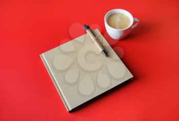 Closed blank book, coffee cup and pen on red paper background.