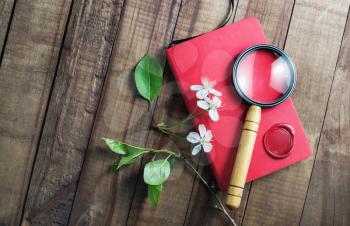 Photo of blank red notebook, magnifier, cherry flowers and green leaves on wooden background. Stationery elements.