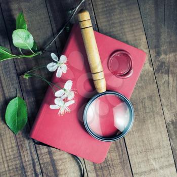 Red notebook, magnifier, wax seal, herry flowers and green leaves on vintage wood table background.