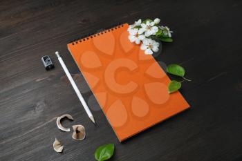 Blank orange notebook, pencil, sharpener and flowers on wooden background.