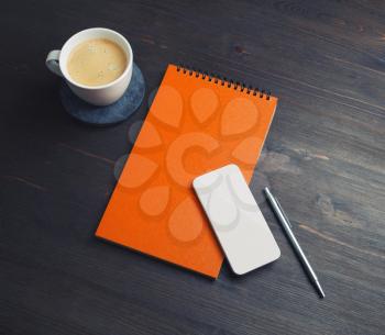 Blank business template. Blank orange notebook, smartphone, coffee cup and pen on wood table background.