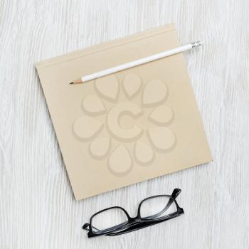 Blank square booklet, glasses and pencil on light wooden background. Template for placing your design. Flat lay.