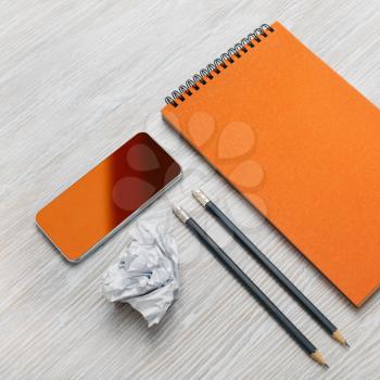 Photo of blank stationery set on light wood table background. Blank orange notebook, smartphone, pencils and crumpled paper.