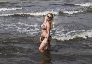 Beautiful blonde standing in sea water. Girl with sunglasses on the sea beach. Rest on the sea beach.