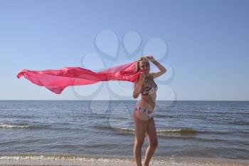 Blond girl in a bikini standing on the beach and holding a red silk handkerchief. Beautiful young woman in a colorful bikini on sea background.